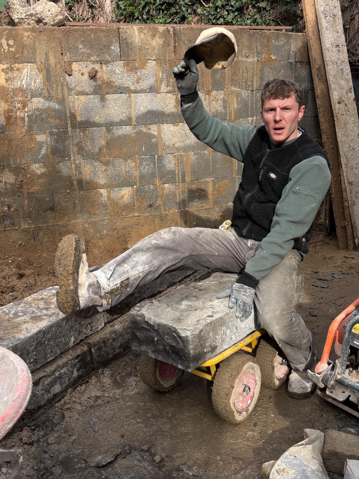 Landscaper resting on a stone slab loaded onto a barrow, cap raised to camera