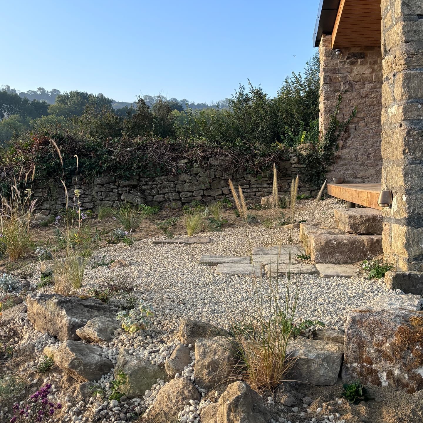 Newly planted gravel garden with limestone boulders and grasses beside a drystone wall, with timber steps up to the garden studio at Studio Garden, Somerset